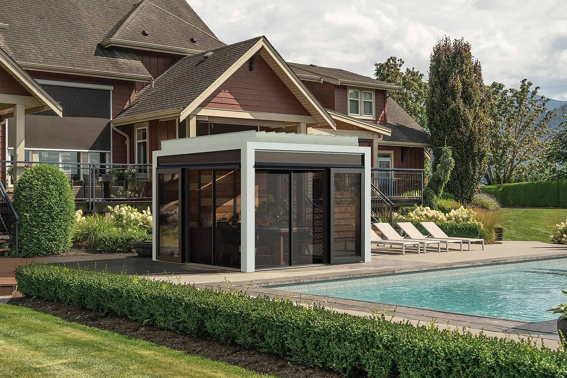 Poolside installation of Stella Fully Enclosed Pergola with a crisp white aluminium frame and transparent black-framed sliding glass doors. Inside, modern patio furniture is visible, arranged around a central dining surface. The surrounding outdoor setting includes a turquoise swimming pool in the foreground, evergreen hedges, and a residential house behind the structure.
