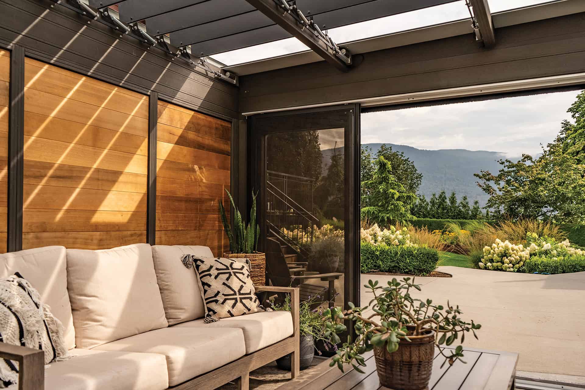 Dark-framed version of Stella Fully Enclosed Pergola set into a landscaped backyard next to a rectangular pool. The structure features floor-to-ceiling glass sliding doors with dark aluminium frames, allowing clear visibility of wooden chairs with green cushions placed inside. The roof louvres are tightly aligned, forming a uniform shaded cover. Surrounding elements include trimmed hedges, a paved pool deck, and a modern house.