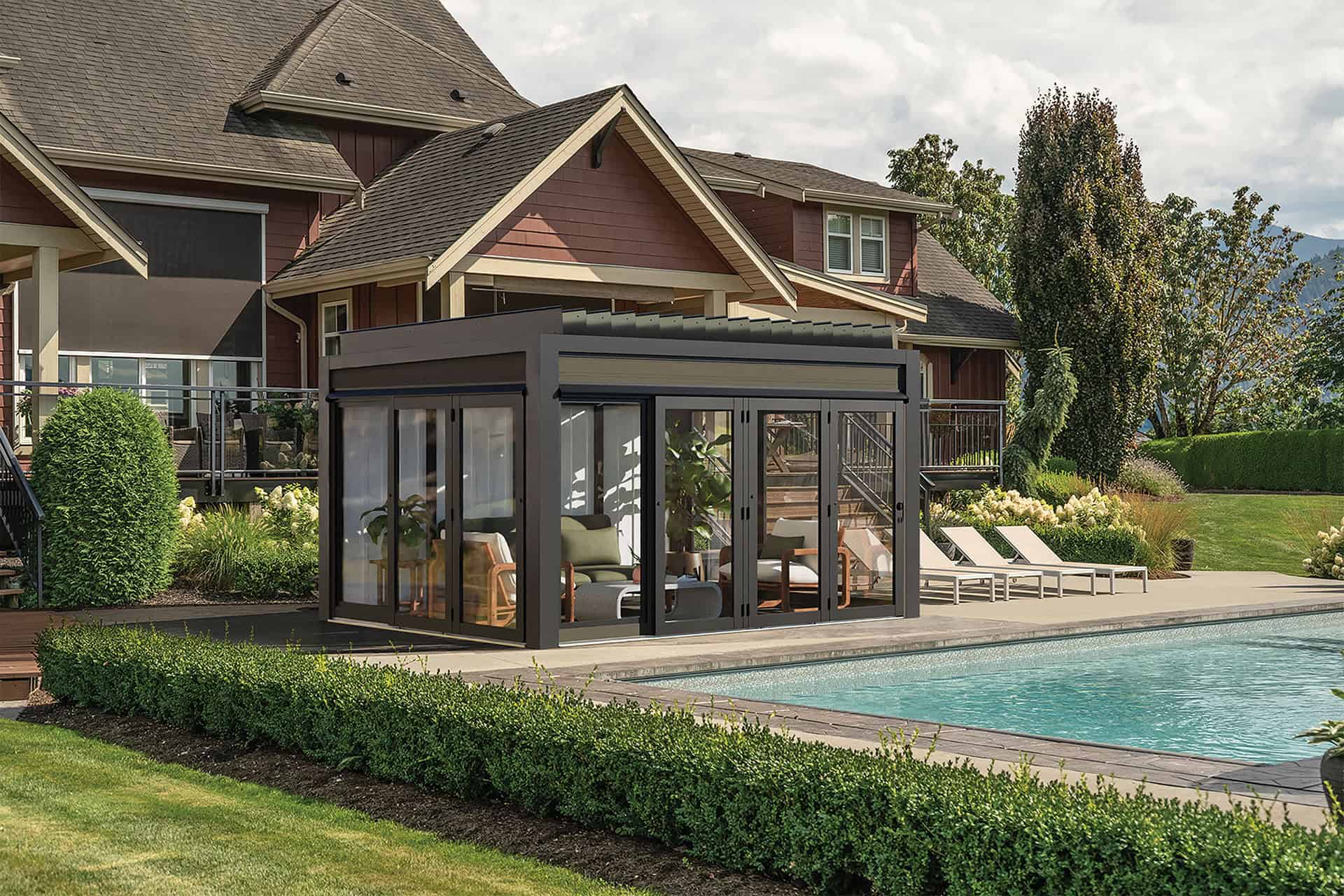 White-framed Stella Fully Enclosed Pergola positioned beside a swimming pool with water reflecting the structure. The pergola is fitted with tall black-framed sliding glass doors, revealing a modern dining table and chairs neatly arranged inside. The surrounding landscape features lush hedges, a trimmed lawn, and a residential home partially visible in the background.