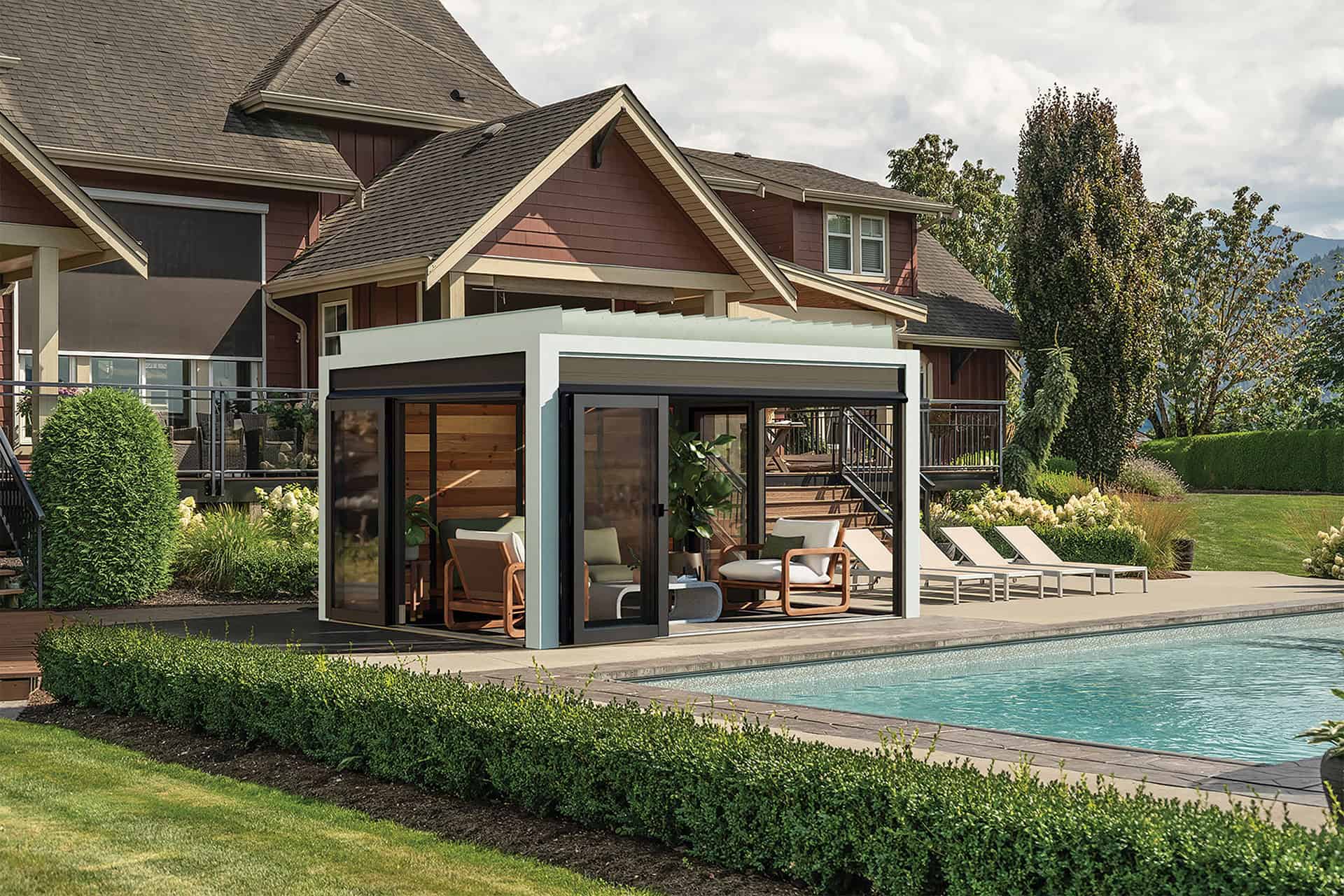 Fully enclosed Stella Pergola installed beside a swimming pool in a landscaped backyard. The pergola features a modern rectangular frame in a light finish with dark trim, full sliding glass doors, and wood panelling inside. Comfortable armchairs and a coffee table are placed within, with greenery and decorative plants visible outside. Four white loungers line the poolside patio in front of the pergola, creating a luxury outdoor living space with manicured hedges, trimmed bushes, and a two-storey house in the background.