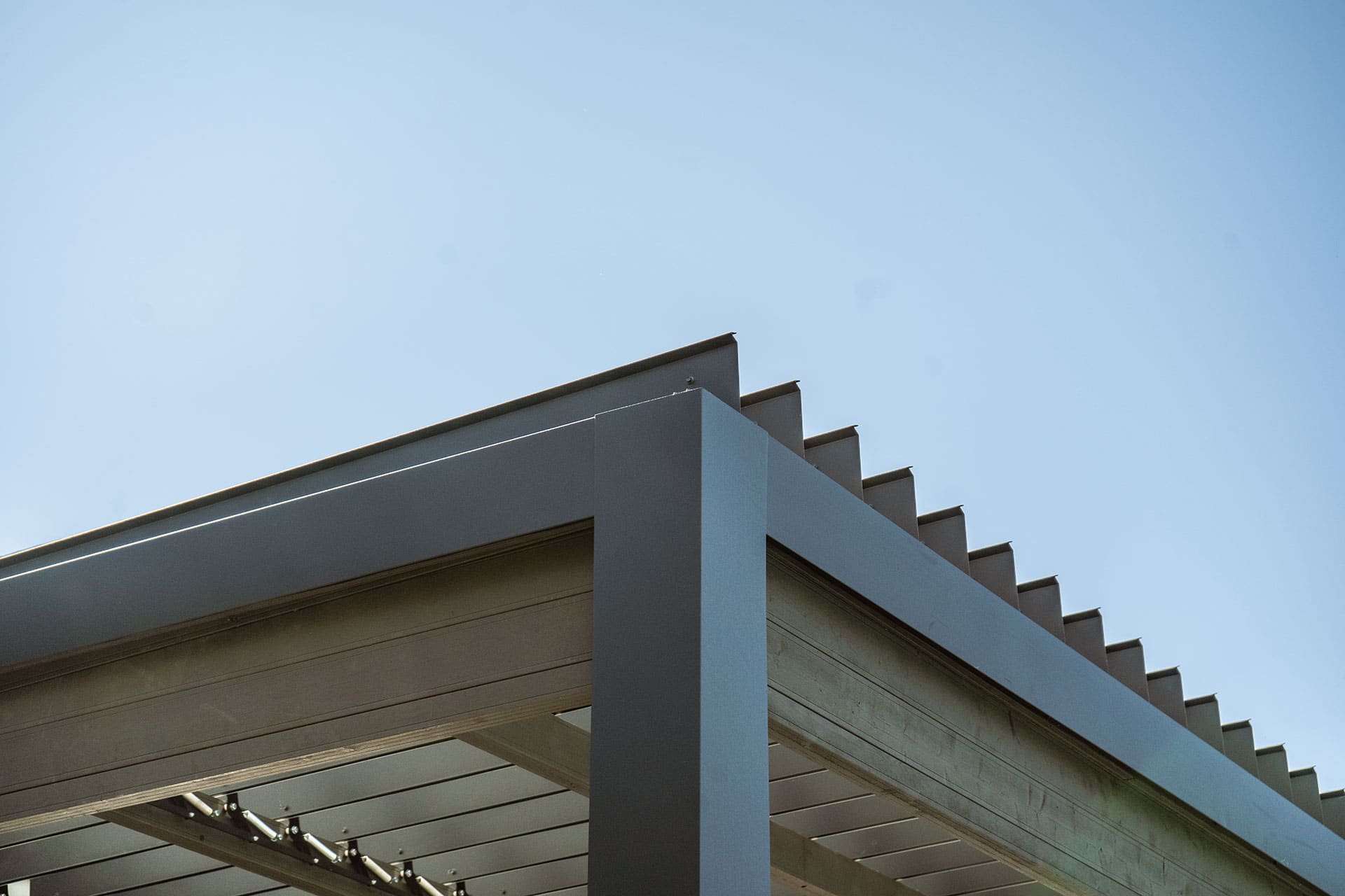 Close-up detail of the Stella Fully Enclosed Pergola’s upper roof edge, showing the clean modern lines of the light-coloured metal frame and overlapping motorised louvres. The angled blades create a layered effect against a clear blue sky, emphasising the sleek architectural design and durable construction of the pergola’s roof.
