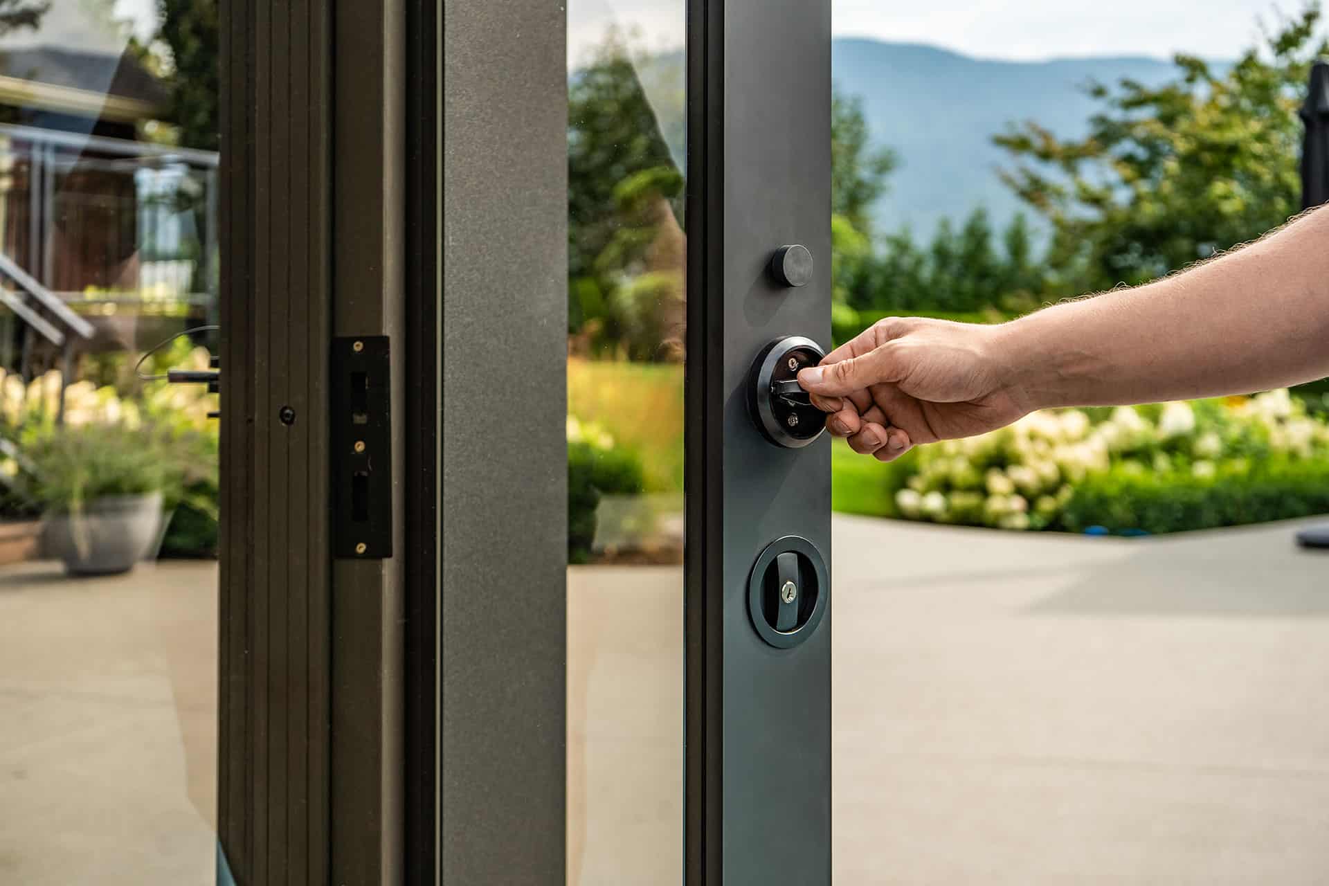 Close-up of a hand locking the Stella Fully Enclosed Pergola’s sliding glass door. The image highlights the modern matte black lock system mounted on the slim aluminium frame, with glass panels reflecting the outdoor garden. The blurred background reveals flowering bushes, a large planter, and distant mountains, showing the security and durability of the pergola’s door system.