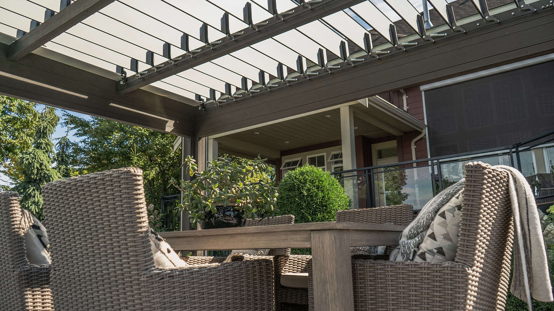 Close-up angled view from beneath the Stella Open Air Pergola, showing the motorised louvre roof system with open slats allowing filtered sunlight. A woven wicker dining set with cushioned chairs and a wooden table is arranged beneath, with a potted plant centred on the tabletop. The backdrop includes part of a red house with a covered patio, white support columns, and manicured landscaping with dense greenery.