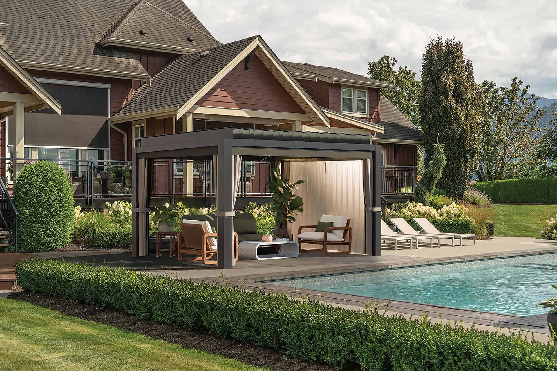 The Stella Open Air Pergola placed by a poolside in a landscaped backyard with a two-storey red house in the background. The dark-framed pergola has partially closed louvres and white side drapes gathered neatly. Inside are cushioned lounge chairs, a sofa, and a low coffee table surrounded by potted plants. To the right of the pergola, several white poolside loungers line the paved patio beside the water.