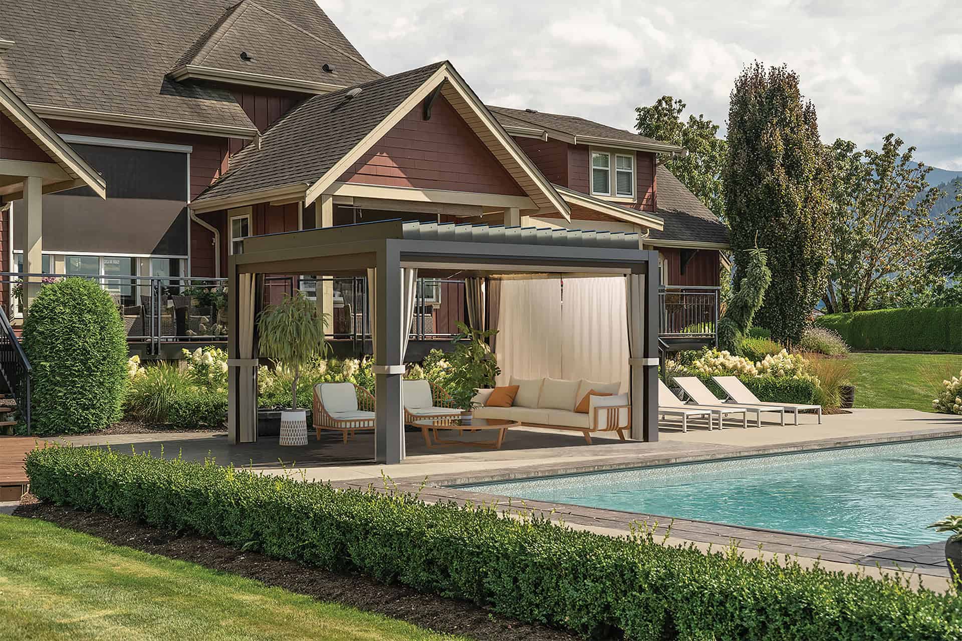 Another poolside view of the Stella Open Air Pergola, showcasing a variation of the furniture inside. This arrangement includes a light sofa with orange accent pillows and two cushioned lounge chairs, all paired with a small side table. The pergola retains its dark aluminium frame and cream side drapes, which are partially drawn. The background features lush hedges, ornamental shrubs, and the pool reflecting the bright daylight.