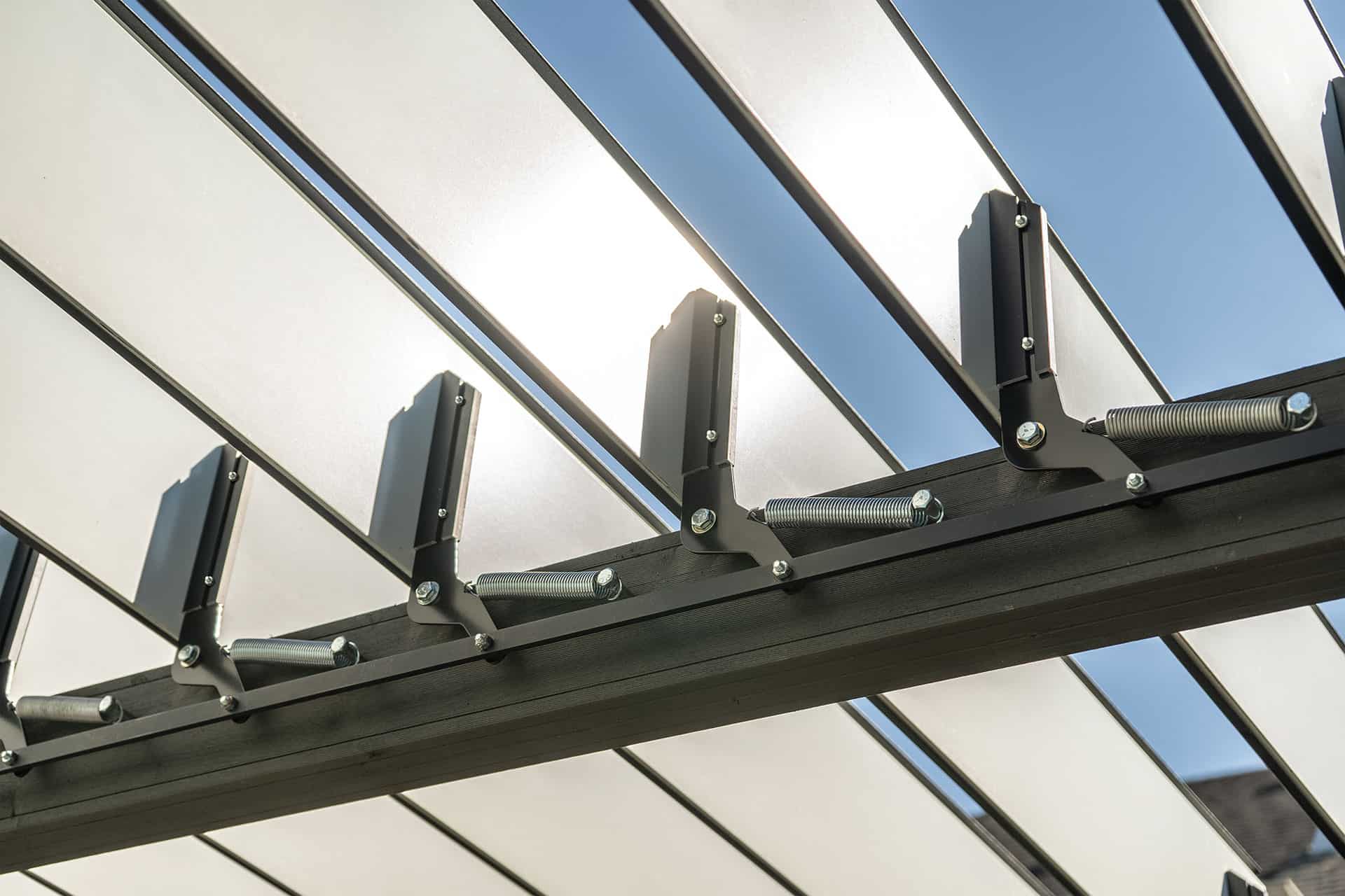 Detailed close-up of the Stella Open Air Pergola roof mechanism, focusing on the black brackets, pivot arms, and tension springs that support the motorised louvres. The metallic components are bolted into the horizontal beam, while the white louvre blades are tilted open above, allowing sunlight to filter through against a clear blue sky.