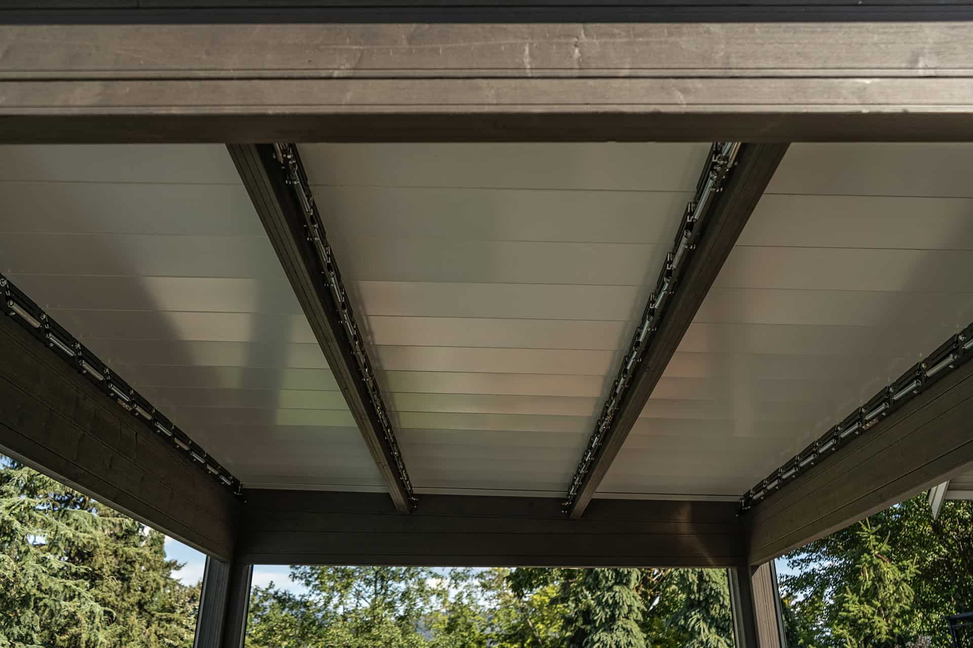 Interior underside view of the Stella Open Air Pergola, showing the louvre system completely closed to form a solid roof. Three main beams run across the width of the pergola, with the motorised louvre panels fitted neatly between them. The finish of the frame is a dark woodgrain-like texture, while tree foliage is visible in the background beneath the closed roof.