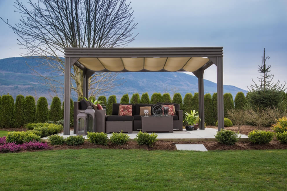Outdoor setting featuring the Valencia Wood Pergola installed on a concrete patio in a landscaped backyard. The pergola has a beige retractable canopy pulled fully across the roof, supported by four dark-wood square posts. Inside, a wicker sectional sofa with black cushions and floral accent pillows surrounds a matching coffee table. Decorative glass lanterns are placed on the left, while potted plants with vibrant green foliage add natural accents to the right side. A row of manicured hedges, a bare tree, and a mountainous horizon line frame the background under a cloudy blue sky.