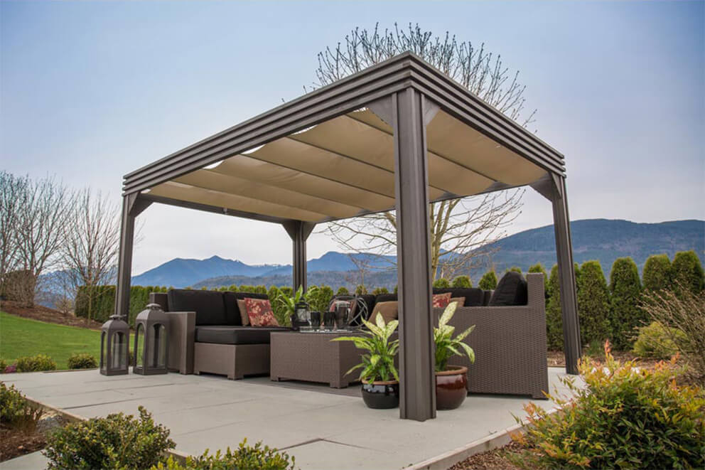 Angled side view of the Valencia Wood Pergola showing the retractable beige canopy fully extended. The structure is positioned on a concrete patio, surrounded by neatly trimmed landscaping with small shrubs and flowering plants. Inside, dark wicker sectional seating with black cushions and patterned pillows is paired with a woven coffee table. Large decorative lanterns stand on the patio’s edge, while potted green plants soften the seating area. Rolling mountains are visible in the background beyond a tall hedge, under a soft cloudy sky.