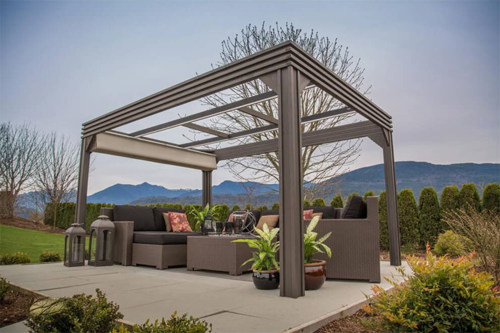 Side angle of the Valencia Wood Pergola with its beige retractable canopy fully retracted, leaving the open dark-wood frame exposed. The square beams and cross-supports of the pergola are clearly visible, showcasing its durable construction. Beneath the structure sits a wicker sectional sofa with black cushions and patterned throw pillows, along with a central coffee table. Potted plants with broad green leaves and two tall black lanterns flank the seating arrangement. Behind the pergola, a line of evergreen hedges stretches across the scene with mountains and overcast sky in the distance.
