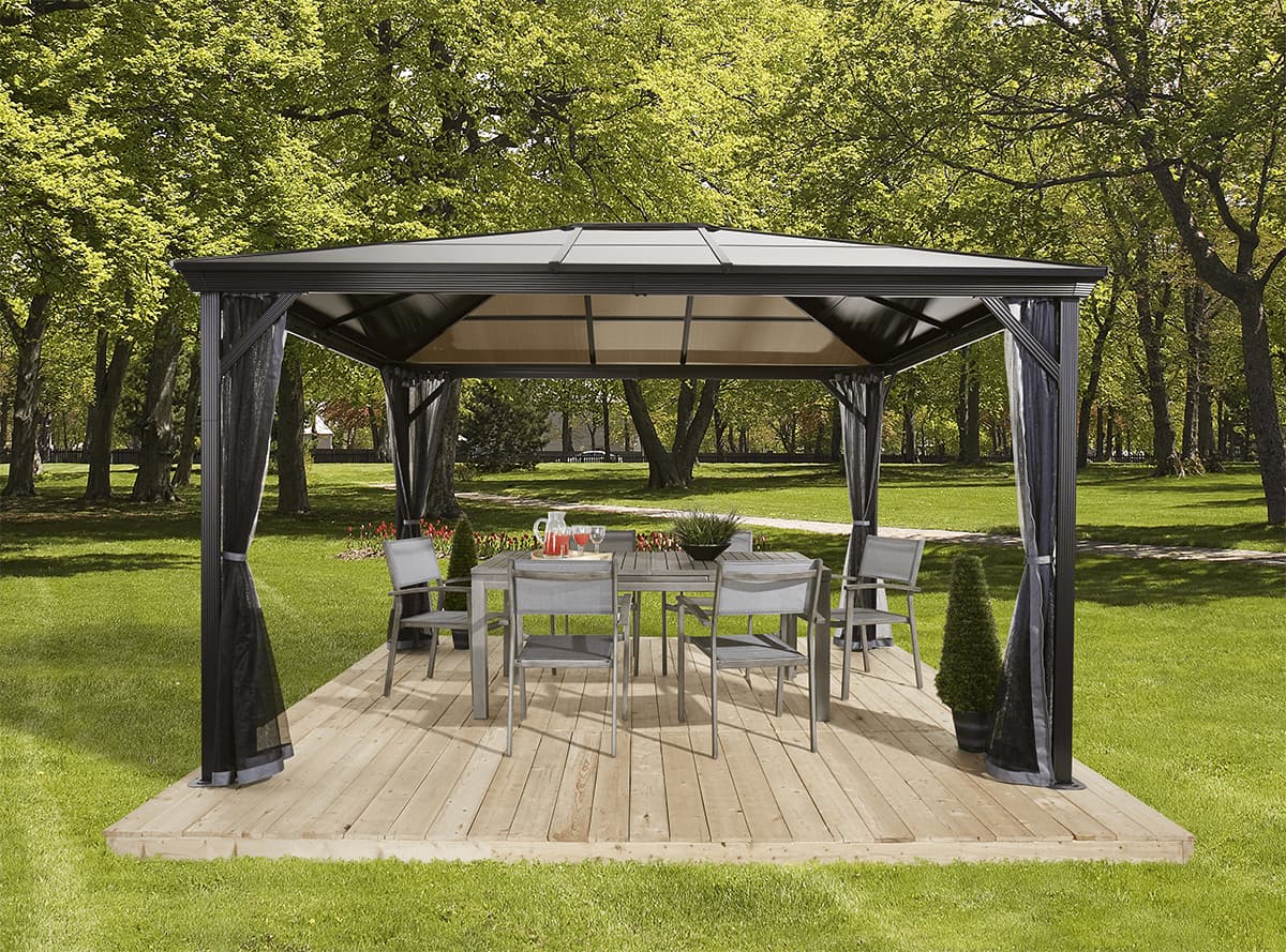 Verona Gazebo staged in a landscaped backyard setting, positioned on a light-coloured wooden deck surrounded by trimmed bushes and a privacy hedge. Beneath the gazebo sits a rectangular grey rattan dining table with six matching dining chairs, cushioned in dark grey fabric. A pitcher of juice and clear glasses are placed on the table, while sheer black mesh curtains are drawn back at each post. Behind the structure, a line of tall evergreen hedges frames the view, and in the distance, rolling mountains rise under a pale blue sky.