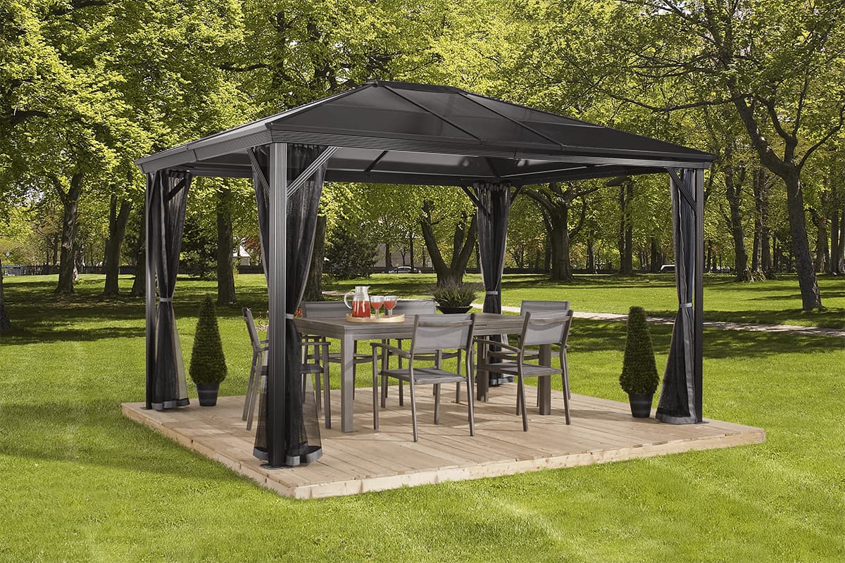Angled left-side view of the Verona Gazebo, placed on a wooden deck in a garden setting. A full outdoor dining set, including a rectangular wicker-style table and six dark-cushioned chairs, sits underneath. The gazebo’s pitched black metal roof provides solid shade overhead. Each of the four aluminium posts features black mosquito netting draped neatly and tied back, while the background reveals trimmed hedges and distant mountain peaks. Decorative garden lanterns stand at the left front corner of the deck, adding detail to the scene.