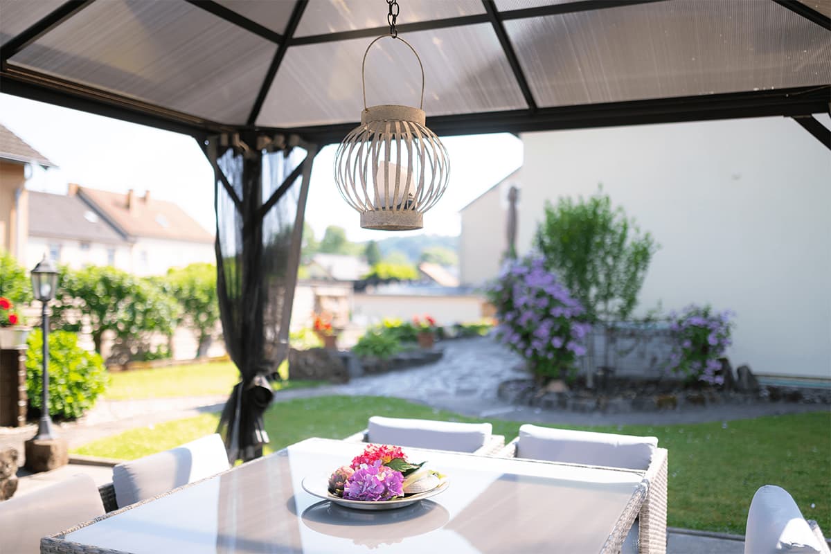Diagonal side view of the Verona Gazebo showing its structure with the retractable mosquito netting open, exposing the interior. The roof’s dark pitched aluminium slats extend outward slightly, creating overhangs. A six-piece rattan dining set with black cushions sits beneath, centred around a rectangular table holding decorative items and glassware. Large black lanterns rest on the deck floor in front, while vibrant green potted plants frame the scene. Behind the gazebo, evergreen hedges and mountain ridges extend across the horizon.
