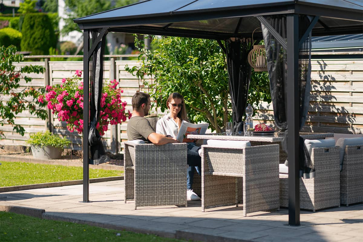 Close-up interior view of the Verona Gazebo, looking upward at the solid black pitched roof with structural beams. A black decorative lantern is suspended from the roof’s central ridge beam, hanging directly above a glossy rectangular dining table with glasses placed on top. The rattan dining chairs surrounding the table have dark grey cushions. In the background, sheer black mosquito mesh curtains hang in loose folds along the sides, with glimpses of flower beds and a stone pathway visible through the opening.