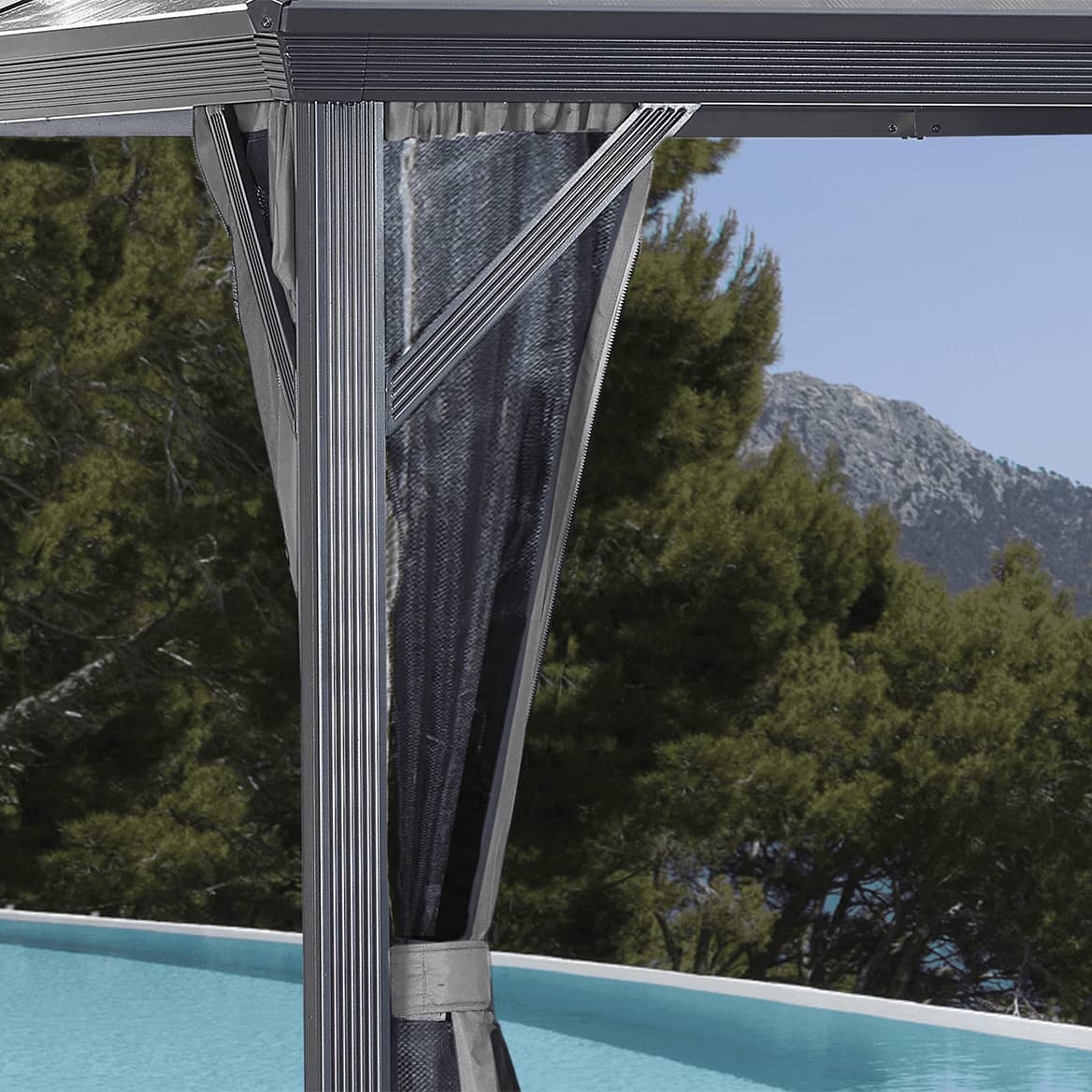 Close-up corner detail of the Verona Gazebo highlighting the aluminium post with vertical ribbing, diagonal roof bracing, and the underside of the sloped black roof. A sheer black mosquito curtain is tied back securely around the post, with fine pleated folds running vertically. In the background, a rattan dining table is visible, topped with a tray holding a clear jug of red juice and glasses. Beyond the gazebo, a landscaped garden with greenery and flowers creates a bright contrast.
