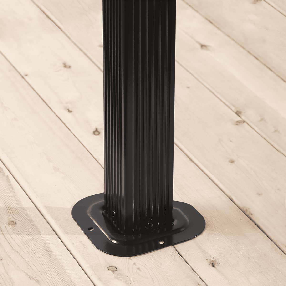 Corner section of the Verona Gazebo positioned in a poolside setting, showing a single ribbed aluminium post, diagonal bracing beam, and part of the pitched black roof. The attached mosquito netting is tied neatly to the post. Behind the gazebo, a vivid blue swimming pool is visible, bordered by tall evergreen trees, with distant mountain scenery completing the background view.