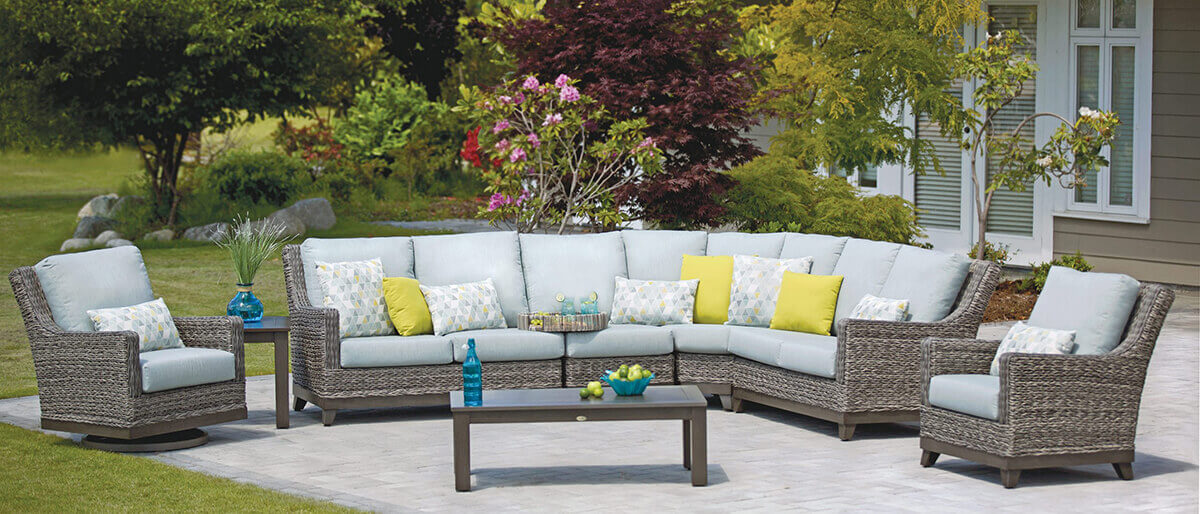 Expansive outdoor sectional layout from the Asheville Furniture Collection placed on a spacious stone patio surrounded by green trees and flowering plants. The sectional features a high-back modular design with resin wicker frames, grey Sunbrella cushions, and multiple accent pillows in yellow and patterned designs. Matching lounge chairs frame each end, complemented by a central rectangular coffee table and side table, creating a stylish and inviting entertainment space.