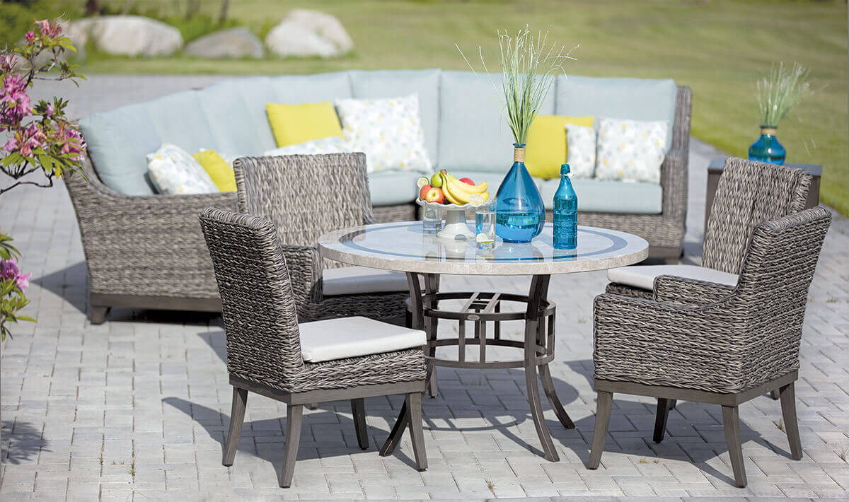 Outdoor dining and seating setup showcasing the Asheville Furniture Collection. In the foreground, a round dining table with a glass top and woven wicker frame is surrounded by four matching dining chairs with cushioned seats. In the background, a modular high-back sectional sofa with grey Sunbrella cushions and colourful accent pillows completes the scene. Decorative blue vases and a fruit bowl add vibrant accents, set against a garden backdrop with bright greenery and flowers.