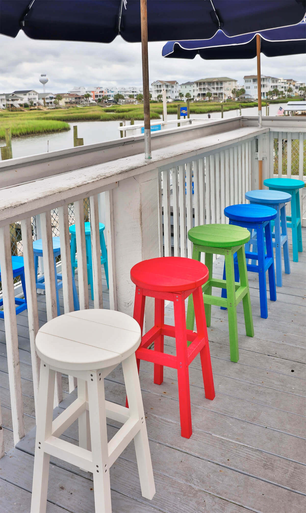 Outdoor boardwalk dining area featuring multiple CRP Bar Collection Backless Bar Stools arranged in a row beneath a navy-blue umbrella. The stools are in a variety of vibrant colours including white, red, green, blue, sky blue, and turquoise. Each stool has a round, backless seat with two wide slats and sturdy, four-legged construction reinforced with square crossbars. They are positioned along a white-painted wooden railing, overlooking a waterfront scene with green marsh grass, a calm body of water, and houses in the background. The setting conveys a casual, coastal atmosphere perfect for gatherings and relaxed outdoor dining.