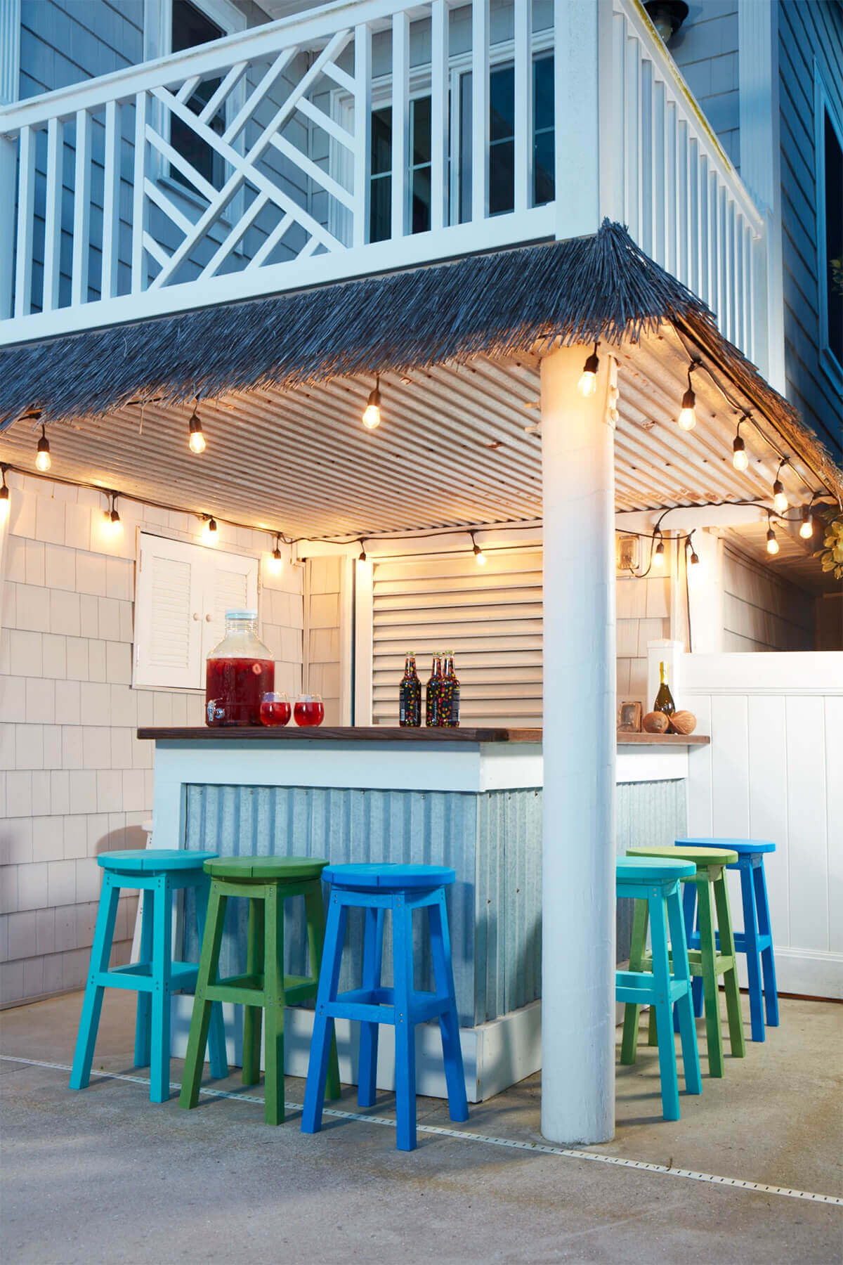 Outdoor tiki-style bar setup at dusk featuring multiple CRP Bar Collection Backless Bar Stools in bright colours, including blue, green, and turquoise. The stools are neatly arranged along the front and side of a corrugated metal bar with a dark wood countertop, under a slanted canopy with thatched roofing. Warm string lights hang above, casting a soft glow across the bar area. A large white support column stands in the foreground. On the counter, a large glass drink dispenser filled with red punch and two glasses sit beside a few decorative bottles, adding a welcoming touch. The surrounding walls are clad in grey cedar shingles with a white balcony railing visible above, creating a relaxed, coastal outdoor entertainment space.