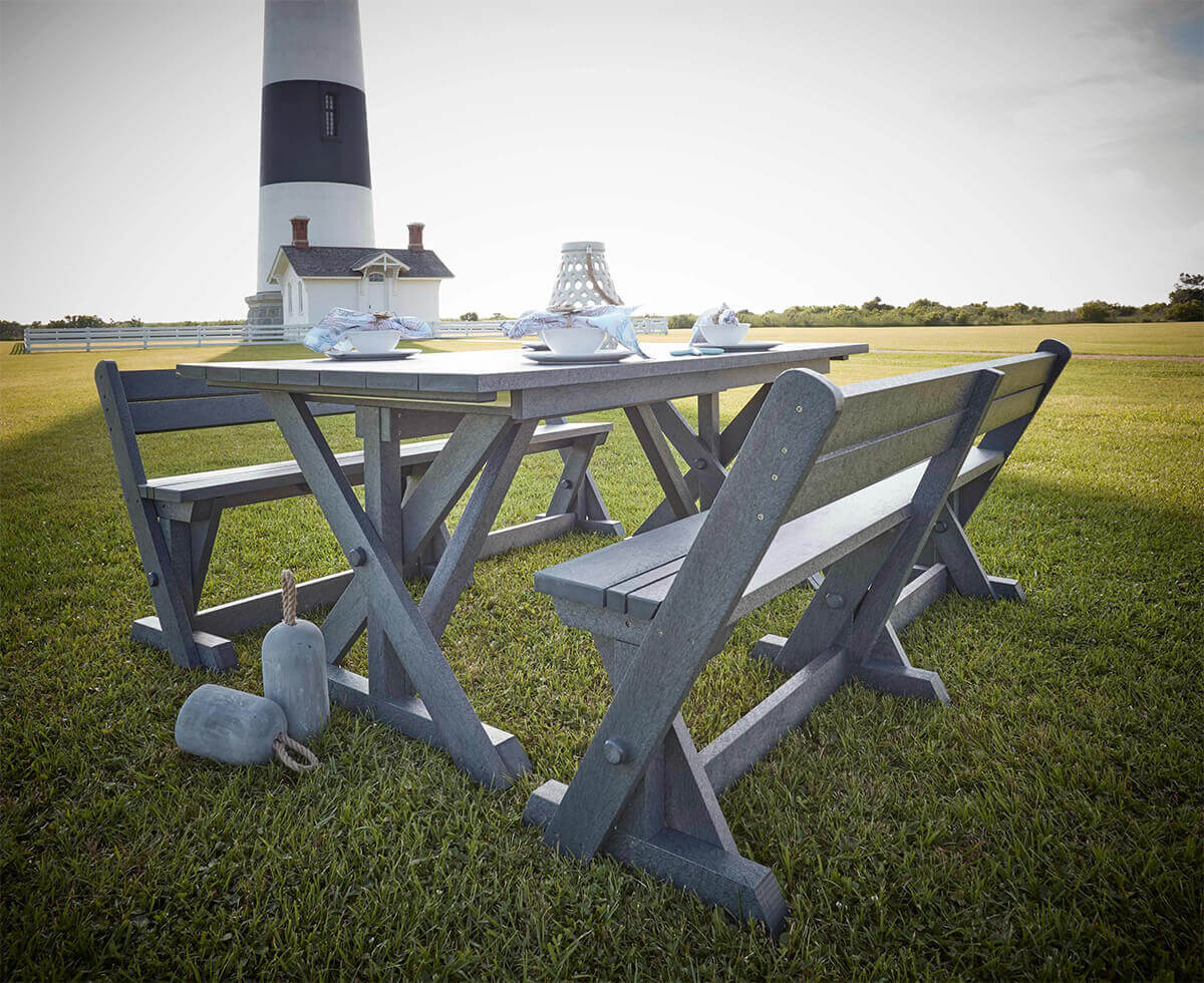 Outdoor setting showcasing the CRP Bar Collection Harvest Wide Dining Table and Harvest Dining Benches in slate grey, placed on a lush green lawn near a lighthouse. The rectangular dining table features a broad plank-style top supported by sturdy trestle-style X-frame legs. Two matching benches with angled legs and supportive crossbars are positioned on each side of the table, creating a rustic, communal arrangement. The table is set with white dishes, bowls, and a decorative lantern, enhancing its welcoming look. Decorative nautical weights with rope handles rest on the grass near the table, adding a coastal theme. The background features a lighthouse with bold black and white stripes, a small outbuilding, and expansive open sky, creating a serene coastal backdrop ideal for gatherings.
