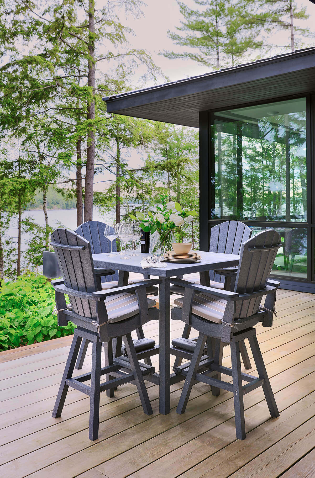Outdoor dining setup featuring the Square Pub Table and four Addy Swivel Dining Arm Chairs from the CRP Bar Collection in slate grey. The square table has a wide, flat top with a central umbrella hole, supported by sturdy, straight legs joined with crossbars for stability. Each matching swivel arm chair has a tall, fan-back design with wide vertical slats, broad armrests, and cushioned striped seat pads secured with ties. The chairs are raised on durable bases with integrated footrests, designed for pub-height comfort. The set is placed on a modern wooden deck overlooking a forested lakeside, blending stylish functionality with a serene natural backdrop.