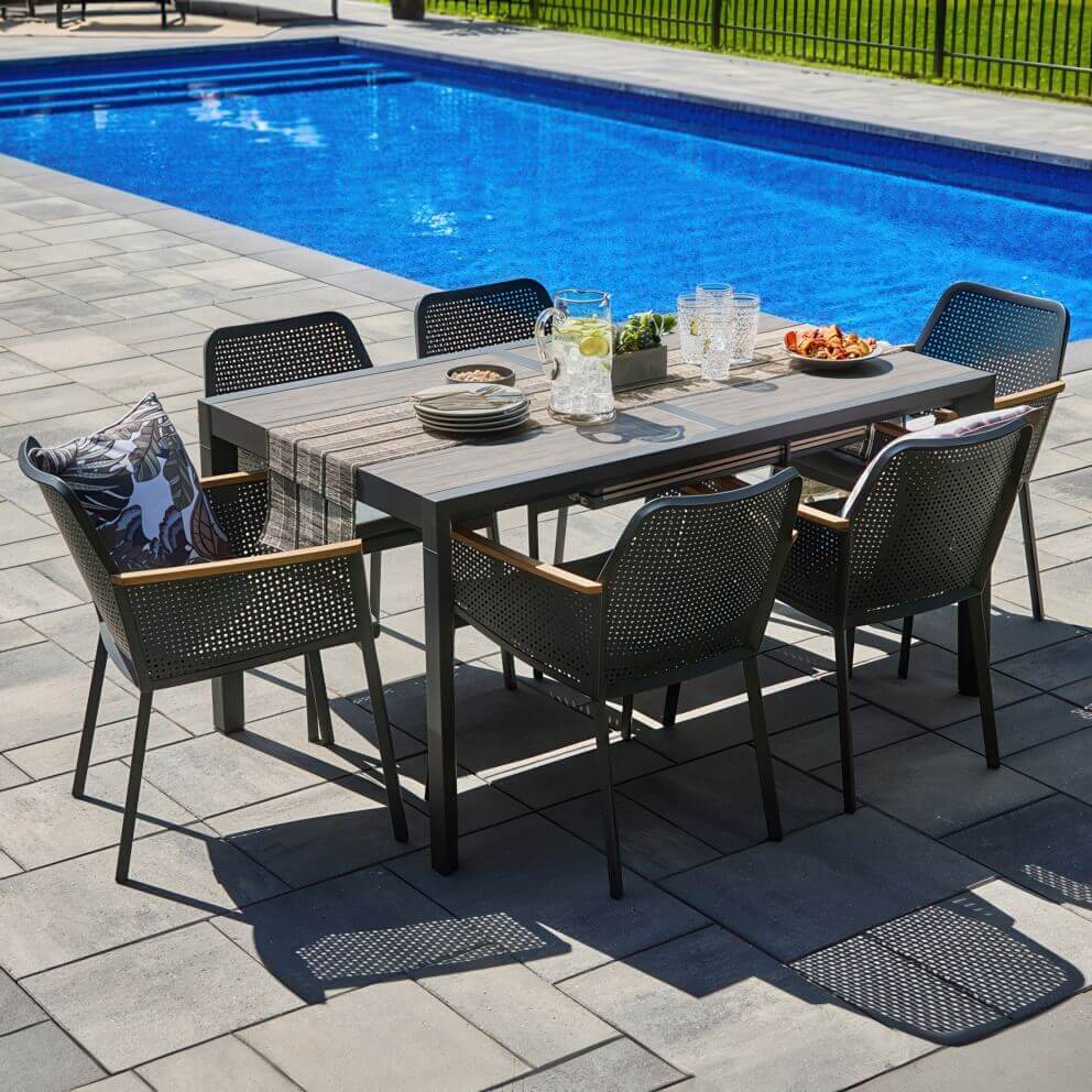 Outdoor Oslo Dining Collection arranged beside a bright blue swimming pool, featuring a sleek rectangular extension table with a grey, wood-look surface and six matching perforated dining chairs with black frames, curved perforated backrests, integrated seat cushions, and light wood-tone armrest inlays. The table is decorated with a woven runner, stacked plates, a jug of water with lemon and lime slices, a bowl of nuts, cutlery, and succulents in a rectangular planter, creating a stylish Scandinavian-inspired patio dining scene.