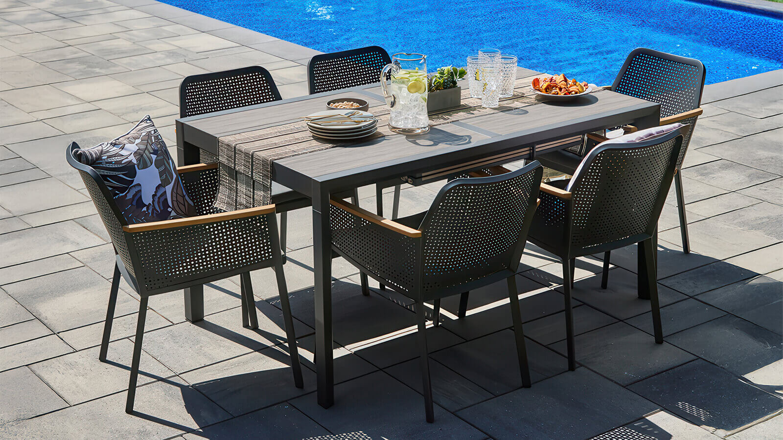 Wide outdoor view of the Oslo Dining Collection placed on a grey stone patio near a shimmering blue pool, showing the full dining set with an extended rectangular table and six perforated black dining chairs. Each chair includes subtle wooden armrest accents and a padded seat cushion. The tabletop is neatly styled with a woven runner, glassware, fresh fruit, and decorative plants, highlighting the Scandinavian-inspired clean lines and modern elegance of the collection.