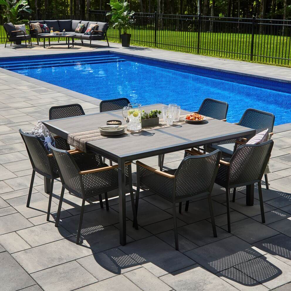 Close angled view of the Oslo Dining Collection table and chairs arranged poolside, focusing on the rectangular extension dining table with a grey slatted-look top framed in black. Surrounding chairs feature a perforated design with smooth curves, wooden armrest accents, and padded dark seat cushions. The table is styled with plates, cutlery, a jug of iced water with citrus, succulents in a planter, and bowls of snacks, showing a modern yet inviting outdoor dining setup.