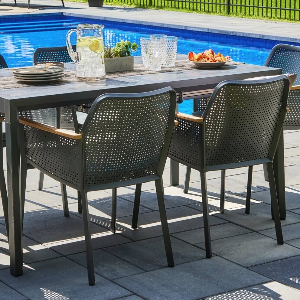 Outdoor Oslo Dining Collection showing the entire eight-seater set with the rectangular extension table and matching perforated black dining chairs, all arranged on a stone patio beside a vibrant blue pool. The table surface is set with a woven runner, crystal glasses, fresh fruit platters, and decorative greenery, while cushions add extra comfort to the chairs. The Scandinavian-inspired design blends sleek black frames, wooden accents, and modern elegance for poolside entertaining.