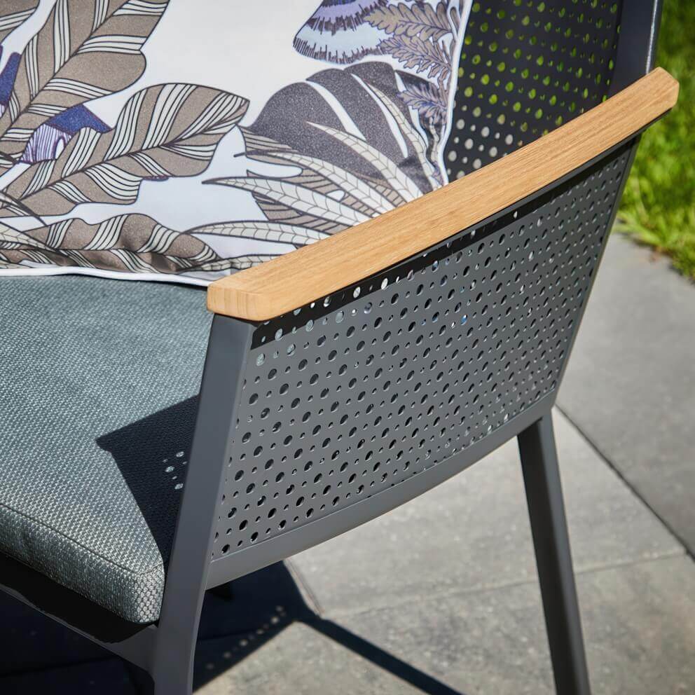 Detail close-up of an Oslo Dining Collection chair in outdoor sunlight, focusing on the perforated black backrest and the smooth, light wood-tone accent running along the top of the armrest. The grey textured seat cushion is visible, with patterned outdoor cushions adding extra style. The perforated design casts intricate shadows on the patio, showcasing the craftsmanship and modern Scandinavian character of the collection.