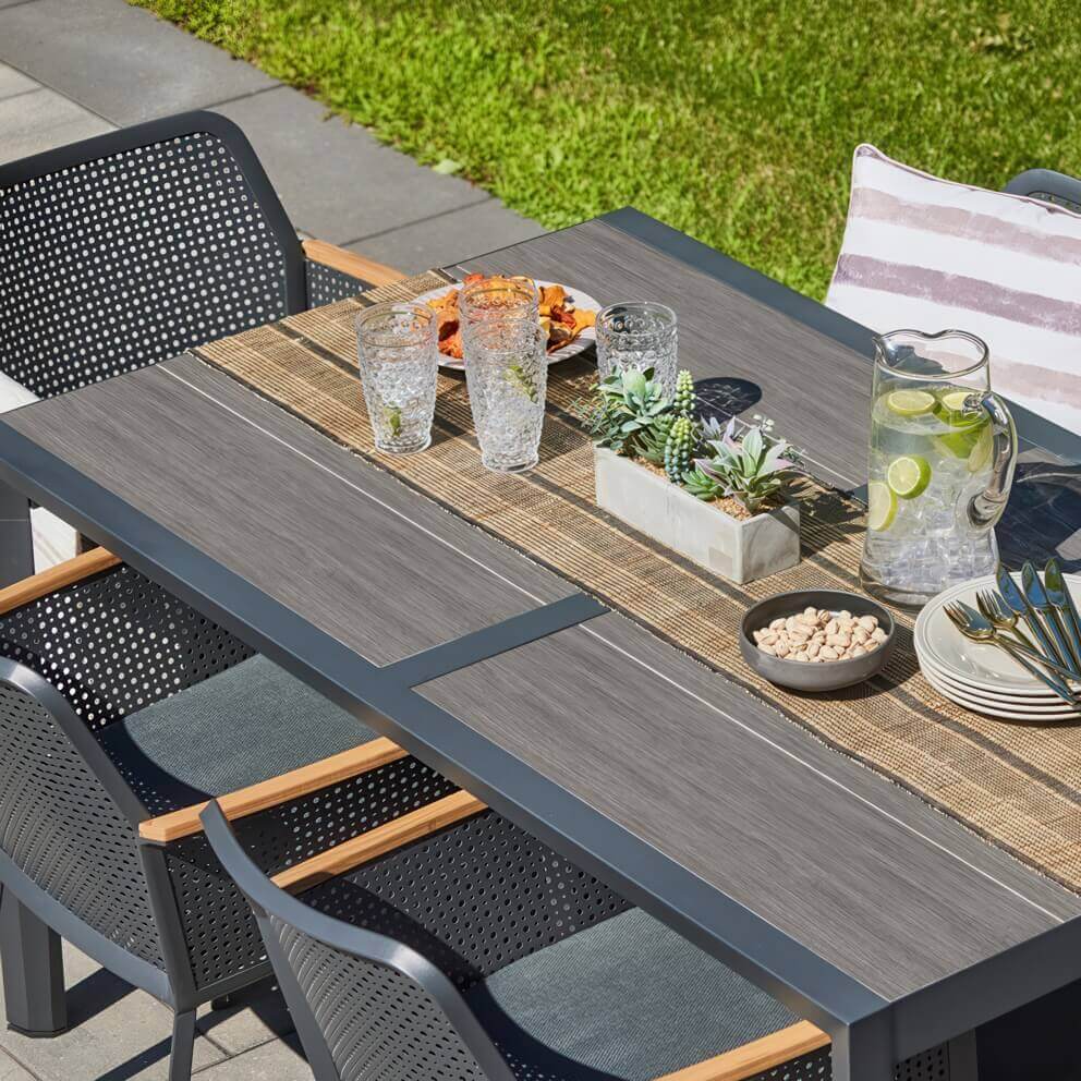 Top-down angled close-up of the Oslo Dining Collection table, highlighting its grey wood-look extension surface framed in black. The table is styled with a woven runner, a glass jug of iced water with citrus, crystal tumblers, plates, gold-toned cutlery, a dish of nuts, and a rectangular planter filled with succulents. The perforated black dining chairs with light wooden arm accents partially frame the table, reflecting the sleek, functional elegance of Scandinavian design.