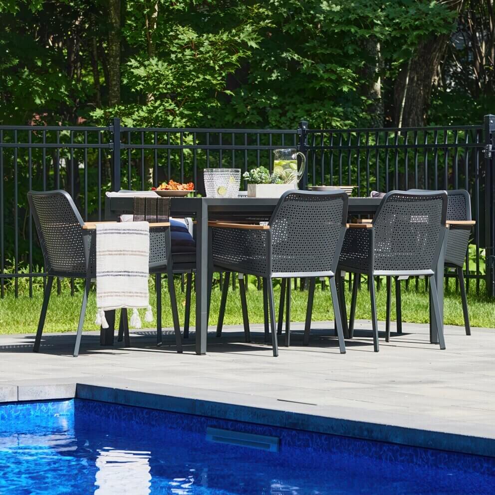 Outdoor scene of the Oslo Dining Collection arranged on a poolside patio, showing the rectangular extension table surrounded by multiple perforated black dining chairs with wooden armrest accents and dark seat cushions. A woven runner with decorative tableware, glassware, and plants sits neatly across the table. In the background, lush greenery contrasts with the sleek, modern lines of the set, reinforcing the Scandinavian-inspired balance of form, comfort, and function.