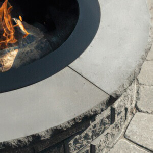Close-up of the StackStone® Fire Pit coping in the Charcoal colour option, showing a dark grey smooth-edged concrete cap encircling the fire pit. The dark coping contrasts with the textured stacked-stone base and the circular black metal fire ring at the centre, which holds burning logs and visible flames. The surrounding patio features interlocking pavers in soft grey tones, enhancing the overall rugged yet refined look.