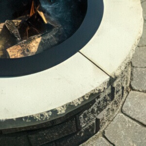 Close-up of the StackStone® Fire Pit coping in the Natural colour option. The coping displays a light beige-grey tone with subtle surface texture and weathered detailing around the edge. It rests atop a multi-toned stone base and surrounds a black metal fire ring containing glowing logs. The soft natural shade complements the neutral patio pavers, giving the fire feature a clean and inviting appearance.