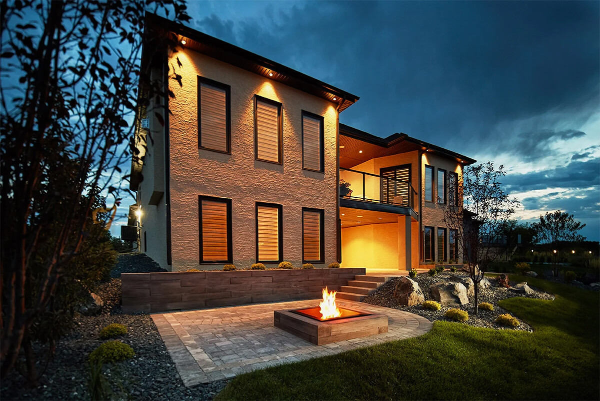 Evening outdoor setting featuring the Bridgewood Fire Pit installed on a light grey brick patio outside a contemporary two storey home. The concrete fire pit is finished in warm cedar brown with an authentic wood grain texture that mimics natural planks, including fine grooves, knots, and tonal streaks. A vibrant flame burns at the centre, enclosed within a black metal spark arrestor. The firelight illuminates the surrounding stonework and modern stucco exterior of the house, reflecting off the smooth glass balcony above. The Look and Feel of Wood in a Concrete Fire Pit, adding warmth and character to a modern outdoor space.