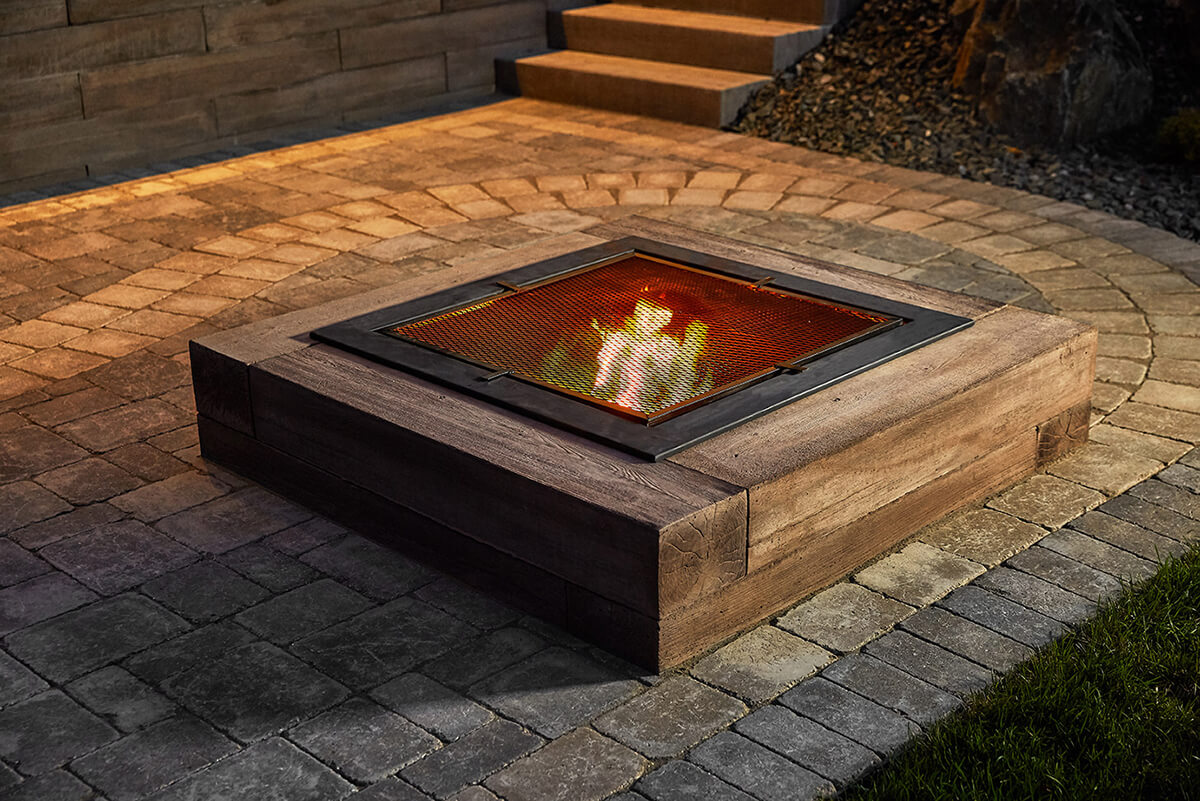 Close up evening view of the Bridgewood Fire Pit glowing on a paved patio. The structure is built from large rectangular concrete blocks designed to resemble aged wood, with visible natural grain, realistic texture, and varied cedar and walnut tones. A black square metal mesh screen covers the flame, which emits a warm orange glow across the rough cobblestone pavers surrounding it. Soft ambient light accentuates the deep wood-like surface detail of the concrete, highlighting its lifelike texture. The Look and Feel of Wood in a Concrete Fire Pit that perfectly blends rustic charm with modern durability.