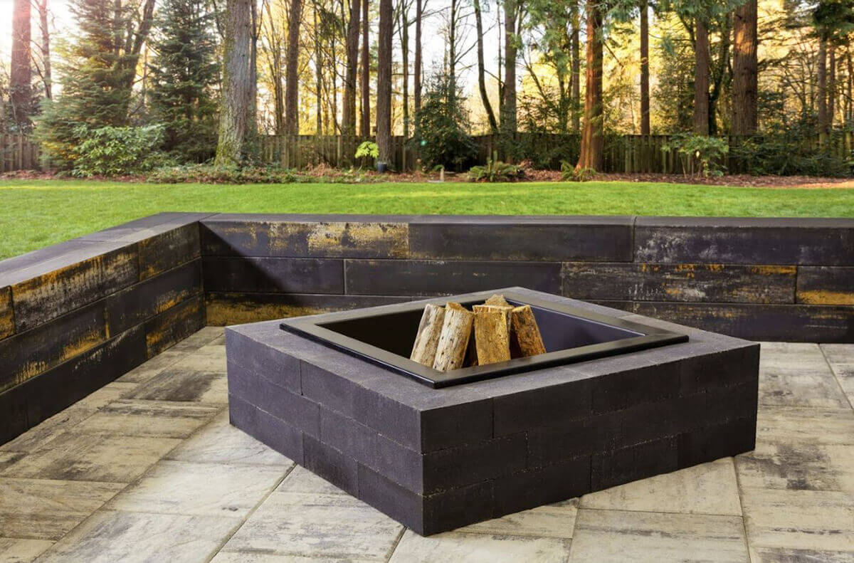 Outdoor patio view of a Caliber Stone Fire Pit surrounded by tall trees and lush greenery. The fire pit is constructed from dark charcoal concrete blocks with a smooth, untextured surface and a square steel insert holding stacked firewood. The pit rests on light grey paving stones bordered by retaining walls featuring golden and rustic streaks. Natural sunlight and dark masonry together enhance its contemporary design. Stylish and ultra modern, add this sleek fire pit to your home for a sophisticated outdoor space.