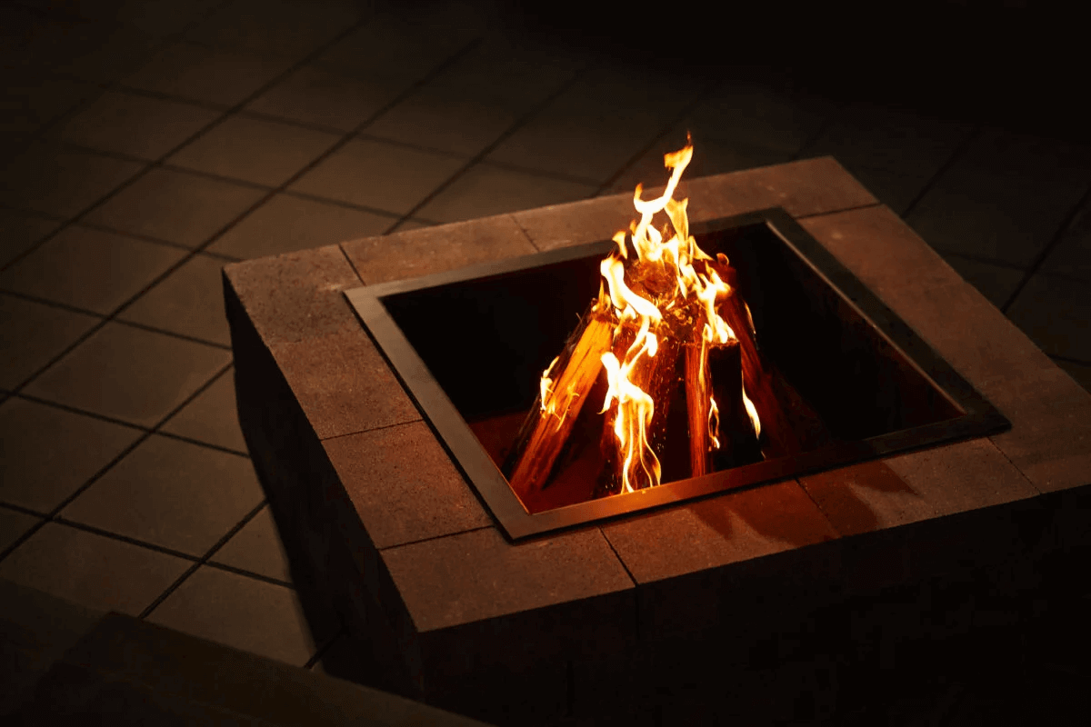 Evening scene showing a Caliber Stone Fire Pit glowing brightly against a dark tiled patio. Flames in vivid yellow and orange rise from firewood inside a matte black steel insert. The surrounding clay brown concrete blocks have clean edges and smooth faces that softly reflect the firelight. The warm tones and minimalist layout create a calm, elegant setting. Stylish and ultra modern, add this sleek fire pit to your home for warmth and visual appeal.