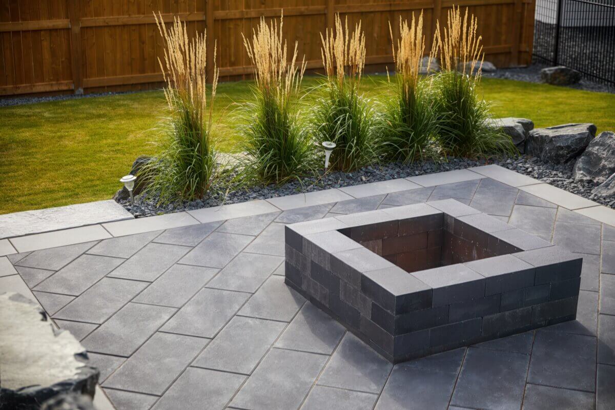 Top view of a Caliber Stone Fire Pit on a spacious grey patio with decorative grasses and green lawn in the background. The fire pit features precisely stacked dark grey concrete blocks with a seamless finish surrounding an empty black steel insert. The geometric symmetry and contrasting textures between stone, grass, and sunlight highlight its modern aesthetic. Stylish and ultra modern, this sleek fire pit completes the look of a contemporary backyard.