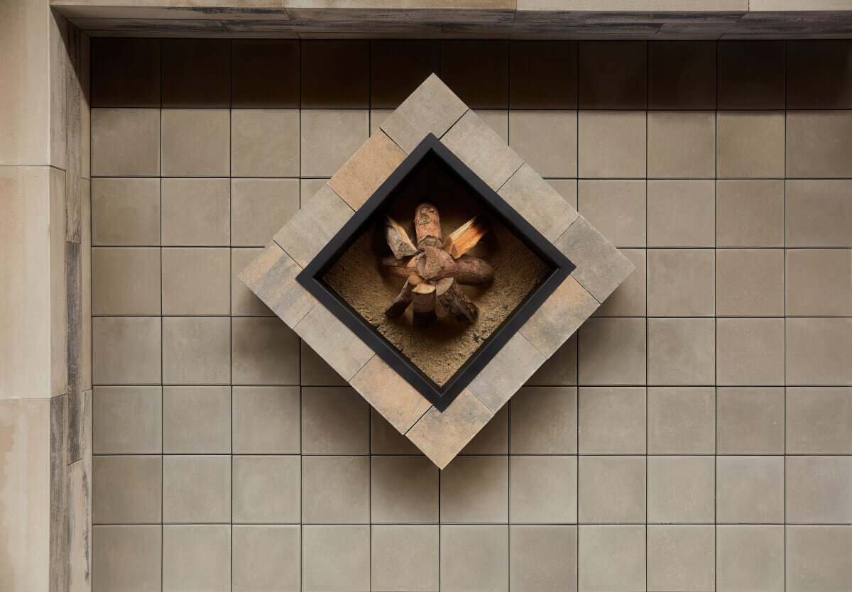 Aerial shot of a Caliber Stone Fire Pit arranged diagonally on a light tan tiled patio. The outer structure is made of smooth concrete blocks in soft taupe and brown shades, surrounding a black steel insert filled with kindling over a sandy base. The clean alignment and warm hues reflect fine craftsmanship and balance. Stylish and ultra modern, add this sleek fire pit to your home for a perfect blend of form and function.
