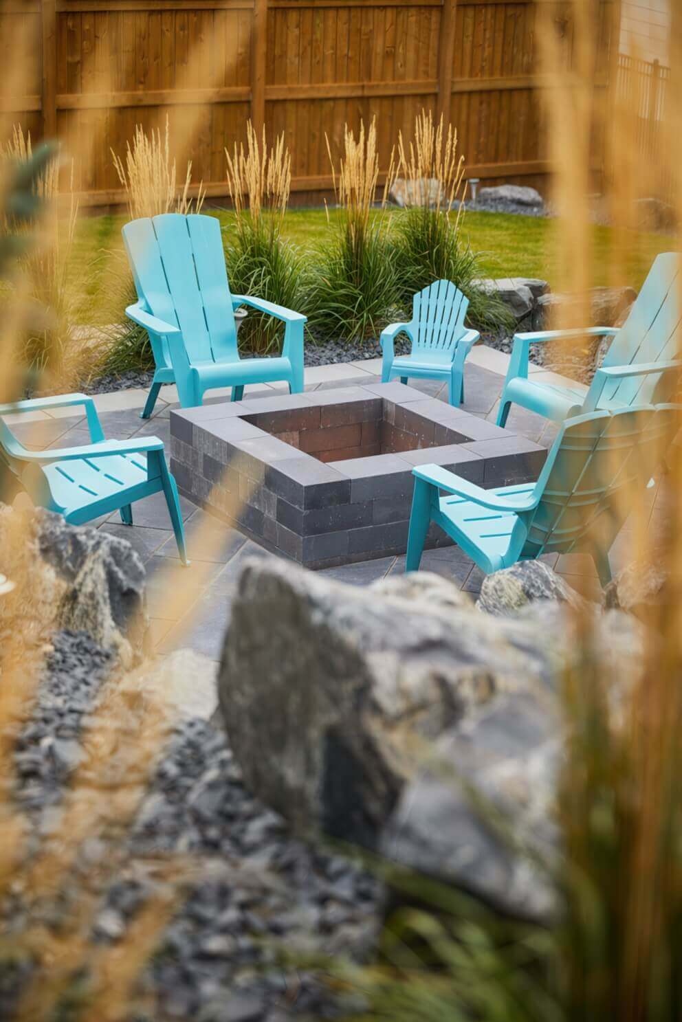 Backyard view featuring a Caliber Stone Fire Pit surrounded by bright turquoise Adirondack chairs. The fire pit consists of dark grey concrete blocks with a matte finish, forming a perfect square shape. Golden ornamental grasses and a wooden fence create a warm contrast with the sleek modern stonework. The clean lines and vibrant setting make this fire pit a beautiful gathering space. Stylish and ultra modern, this sleek fire pit enhances any outdoor retreat.