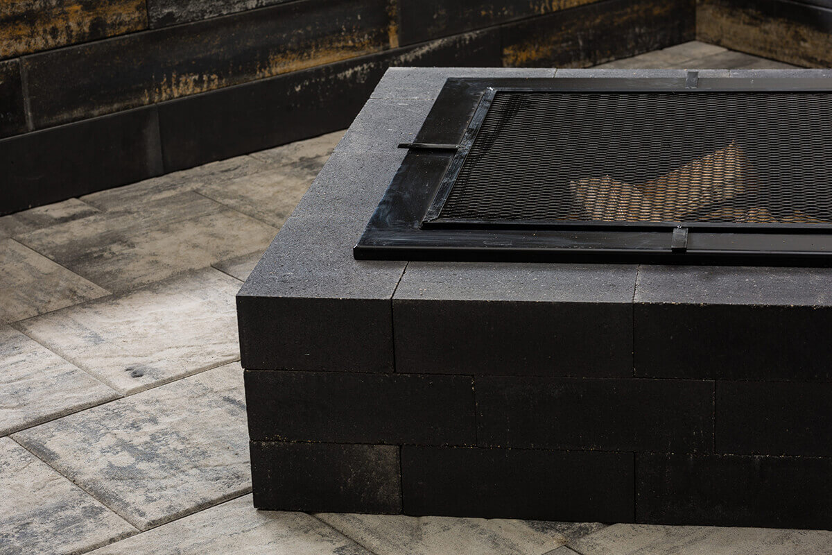 Close up view of the Caliber Stone Fire Pit fitted with a black metal mesh cover. The dark charcoal concrete blocks have a fine smooth finish, and the mesh lid rests securely above the opening. The fire pit is placed on a light grey patio with softly marbled tiles that complement its modern tone. Stylish and ultra modern, add this sleek fire pit to your home for safety and design harmony.