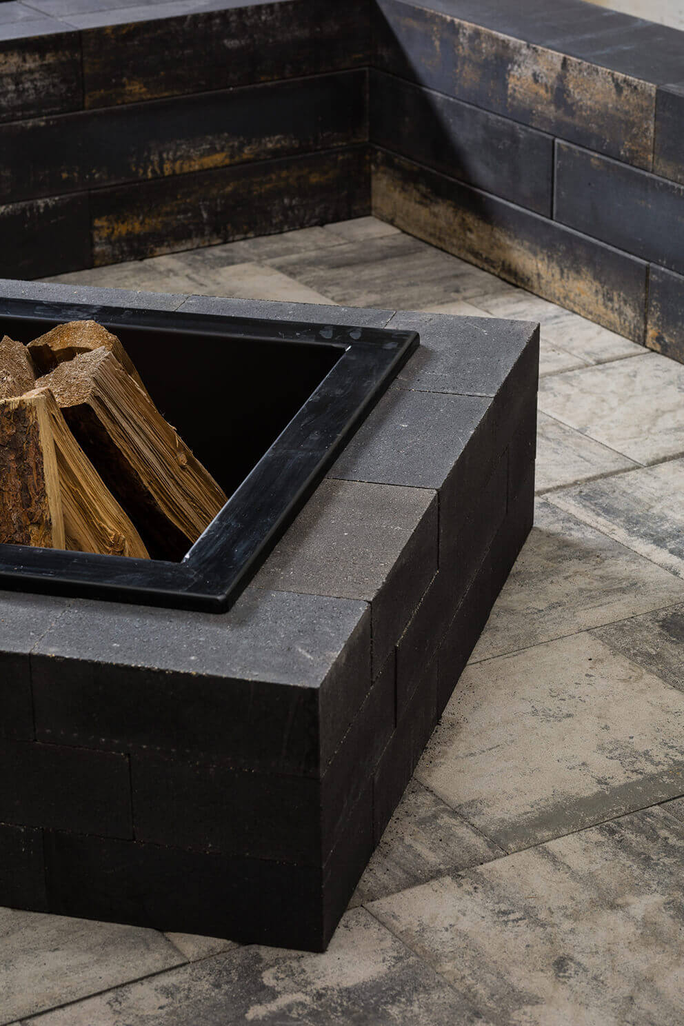 Detailed corner angle of the Caliber Stone Fire Pit showing precise alignment of smooth dark grey blocks with clean rectangular edges. Inside the square pit, a black steel insert holds stacked logs of firewood. The polished surface contrasts beautifully with the natural wood grain and soft lighting. Stylish and ultra modern, this sleek fire pit brings structure and warmth to contemporary outdoor spaces.
