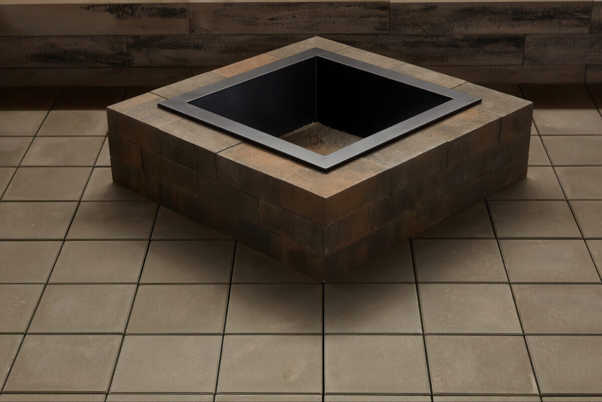 Overhead view of a Caliber Stone Fire Pit placed diagonally on a tan tiled floor. The pit’s structure is made of soft brown and grey smooth concrete blocks surrounding a matte black steel insert with a sandy base. The lighting emphasises the warmth of the natural stone tones and the sleek finish of the design. Stylish and ultra modern, add this sleek fire pit to your home for a clean and elegant touch.
