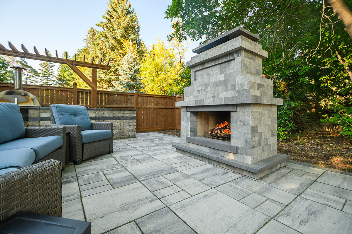 Modern backyard patio featuring the Caliber Stone Fireplace, framed by a tall wooden fence and shaded by lush green trees. The fireplace’s sleek profile is built from smooth grey blocks with deep charcoal accents, complementing the matching stone patio beneath. Flames flicker behind the open hearth, casting a warm glow that enhances the inviting seating area with blue-cushioned wicker chairs. This fireplace reflects the essence of one kit built your way, combining contemporary design with flexible installation.
