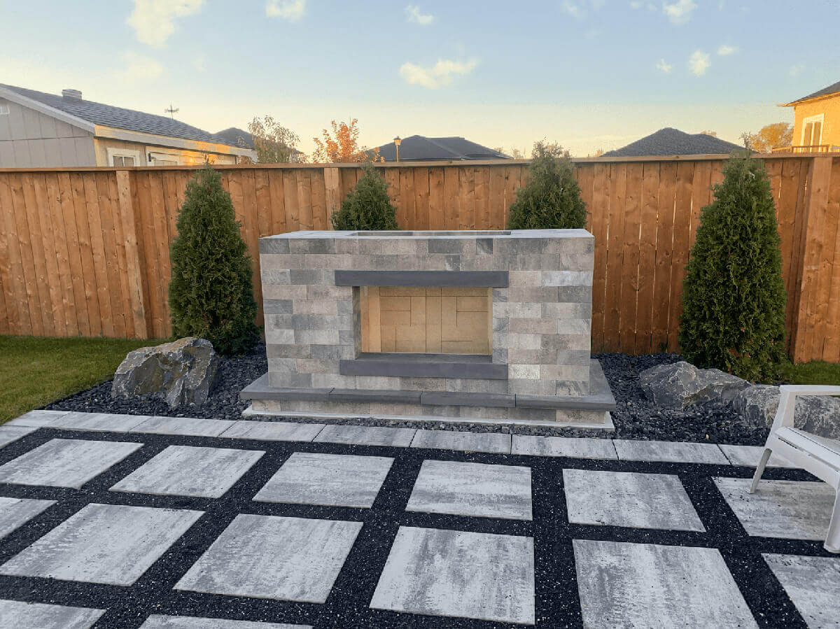 Front-facing view of the Caliber Stone Fireplace set against a cedar privacy fence and trimmed evergreens. The structure, made from precisely cut Sterling grey stone blocks with charcoal trim, sits on a dark stone base surrounded by square pavers arranged in a grid pattern with black gravel joints. The firebox is empty, revealing a light tan interior ready for use. Its clean lines and adaptable layout highlight the Caliber system’s concept of one kit built your way, ideal for modern outdoor settings.