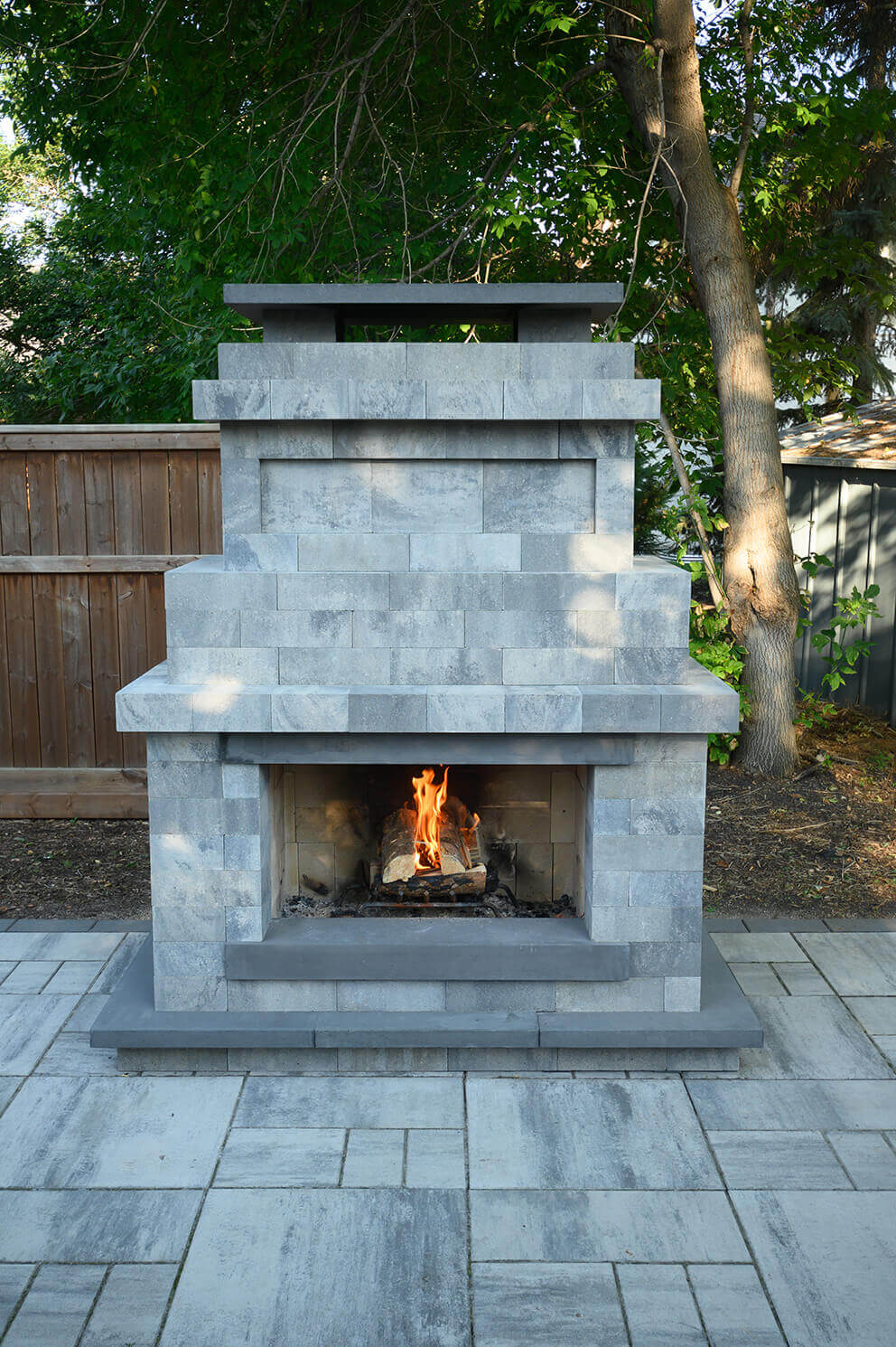Close front view of the Caliber Stone Fireplace, with its tiered, modern silhouette built from pale grey blocks that vary in shade for a natural stone effect. A soft orange flame rises from the central firebox, framed by a dark charcoal mantel and base. The stepped upper tiers create architectural depth beneath a flat chimney cap. Surrounded by trees and bordered by a wooden fence, this design exemplifies the freedom of one kit built your way, merging elegance with practicality.