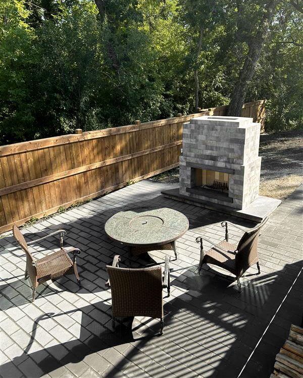 Outdoor patio scene showing the Caliber Stone Fireplace positioned beside a round stone table and four brown wicker chairs. The fireplace stands on grey interlocking pavers enclosed by a wooden fence, with trees providing gentle afternoon shade. Its pale grey stone finish and deep charcoal trim reflect refined craftsmanship. This setup embodies one kit built your way, creating a warm, tailored focal point perfect for social gatherings and relaxed evenings outdoors.