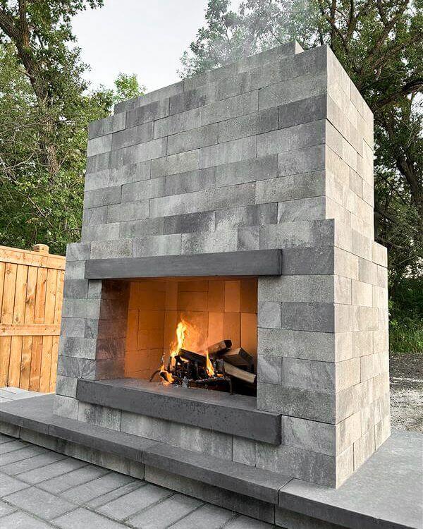 Detailed side view of the Caliber Stone Fireplace, highlighting the sharp geometry of its pale grey Sterling blocks paired with sleek dark charcoal mantel edges. A small controlled fire burns inside, illuminating the light beige firebrick lining. The clean-cut modern design balances strength and simplicity, demonstrating the adaptability of one kit built your way to complement any contemporary landscape.